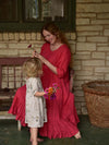 A woman in a red linen dress with 3/4 length sleeves stands and holds a flower, interacting with a young child who is wearing a white floral dress. Angie Dress 3/4 sleeve in Linen